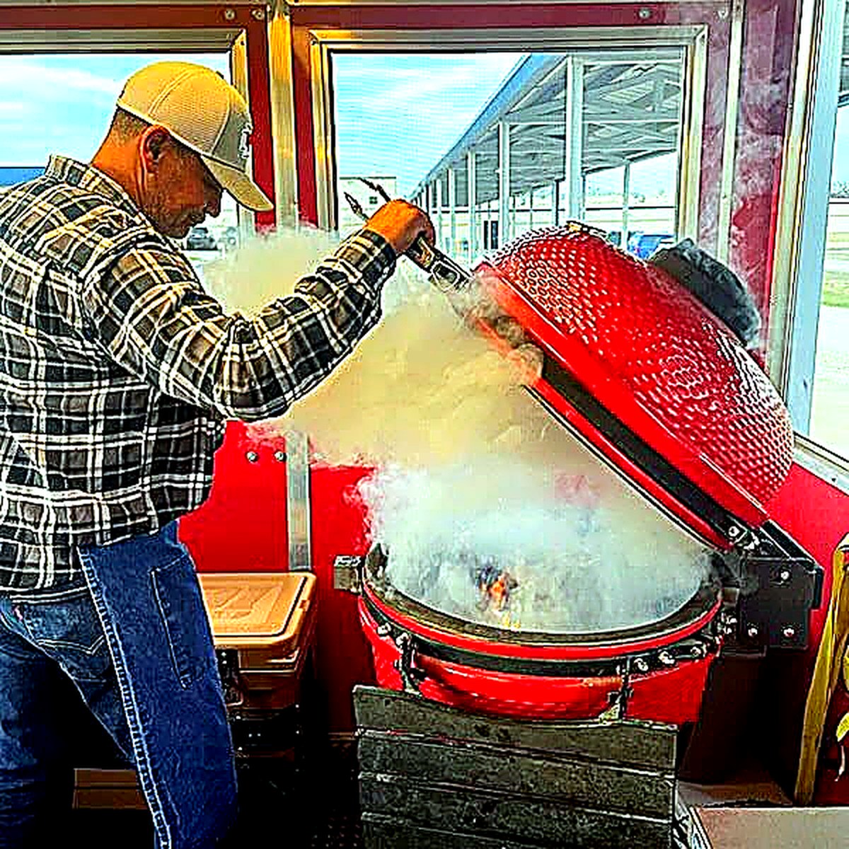 Pitmaster opening a red ceramic smoker with clouds of smoke rising inside the BBQ trailer.